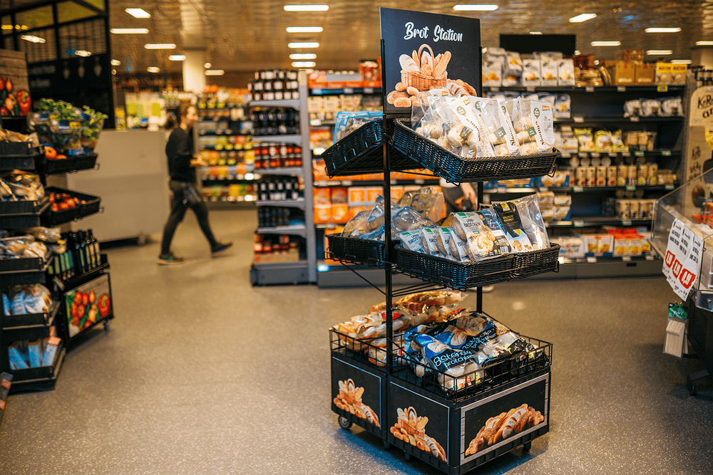 Deuxième point de vente double face avec 6 étagères et un topper (inscription Brot Station, illustration de la corbeille à pain avec diverses pâtisseries), différents pains à cuire sont présentés sur les 6 étagères. Le client est visible en arrière-plan.